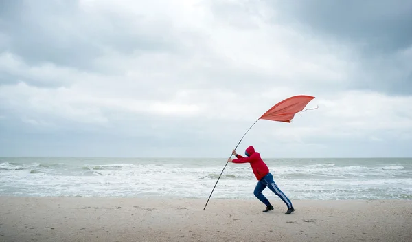 Am Ufer stürmischer See kämpft ein Mensch mit dem Wind. die rote Fahne zeigt die Stärke des Windes an. — Stockfoto