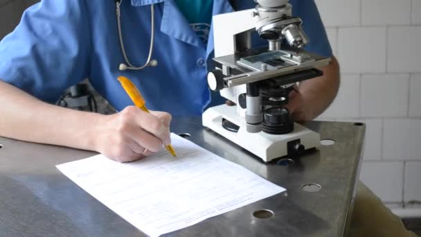 Doctor mirando al microscopio y escribiendo en papel — Vídeo de stock