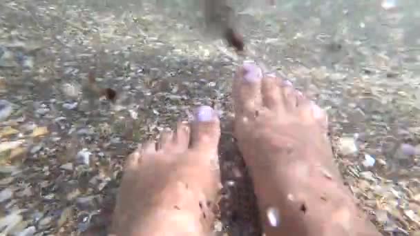 Dos pies femeninos están en agua limpia y transparente en la playa de arena — Vídeo de stock