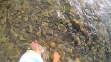 Sığ nehir taşlarının üzerinde çıplak ayakla yürüyen bir kız. Yakın, güneşli bir yaz günü.