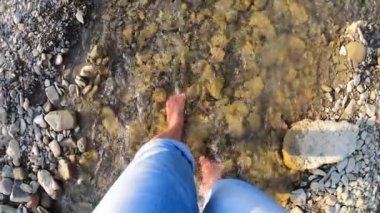 Sığ nehir taşlarının üzerinde çıplak ayakla yürüyen bir kız. Yakın, güneşli bir yaz günü.