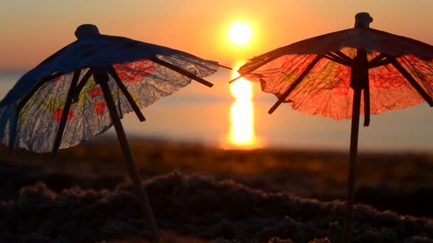 Ombrelli da cocktail di carta in sabbia in riva al mare all'alba del tramonto primo piano. — Video Stock