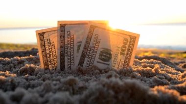 Üç dolarlık banknotlar gün batımında deniz kenarında kumlara gömülür.