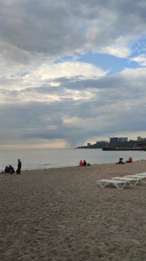 Odessa, Ukrayna, 15 Temmuz 2025, bulutlu bir yaz gününde kumsalda dinlenen ve yürüyen insanlar. Şehir kıyısı yakınlarında ufukta yağmur yağıyor. Deniz manzarası