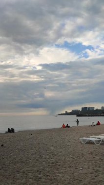 Odessa, Ukrayna, 15 Temmuz 2025, bulutlu bir yaz gününde kumsalda dinlenen ve yürüyen insanlar. Şehir kıyısı yakınlarında ufukta yağmur yağıyor. Deniz manzarası