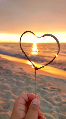 Gündoğumunda, kalp şeklinde yanan maytaplar denizin arka planına karşı. Gün batımında denizde yanan kalp şeklinde maytap. Deniz kıyısındaki bir kadında kalp silueti. Kıvılcımlar. Dikey