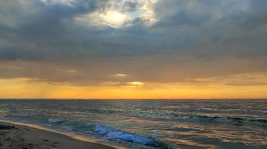 Gün batımında ve gündoğumunda kumsalda mavi suyla deniz dalgaları. Deniz dalgalarının yüzeyindeki güneş ışığının sarı yansıması. Koyu yağmur bulutlarıyla turuncu gökyüzü, ufuk çizgisi ve kumdaki deniz dalgaları..