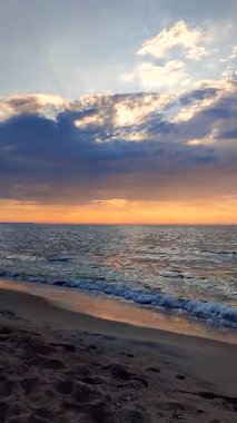 Deniz Burnu. Mavi turuncu gökyüzünde kara yağmurlu bulutlar. Bulutların arkasından gelen güneş ışınları. Gün batımında deniz kıyılarında deniz dalgaları. Deniz dalgalarının yüzeyindeki güneş ışığının sarı yansıması. Dikey