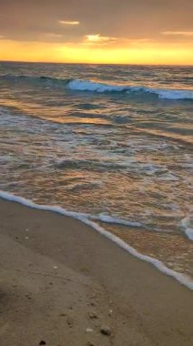 Gün batımında ve gündoğumunda deniz kıyısında mavi şeffaf suyla deniz dalgaları. Deniz dalgalarının yüzeyindeki güneş ışığının sarı yansıması. Bulutlu gökyüzü, ufuk çizgisi ve kumdaki deniz dalgaları. Dikey