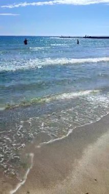 Güneşli, aydınlık bir yaz gününde denizdeki insanlar. Deniz dalgaları, kum, kumlu sahil, deniz kıyısı ve parlak güneş. Güneş deniz dalgalarının yüzeyinde parlıyor. Dikey