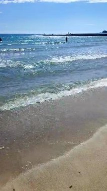 Güneşli, aydınlık bir yaz gününde denizdeki insanlar. Deniz dalgaları, kum, kumlu sahil, deniz kıyısı ve parlak güneş. Güneş deniz dalgalarının yüzeyinde parlıyor. Dikey