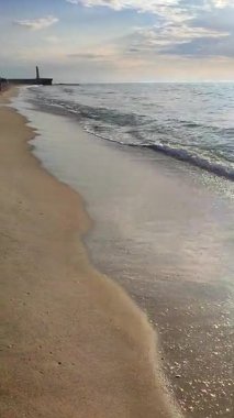 Güneşli bir yaz gününde deniz manzarası. Deniz kıyısındaki kumlu kumsalda dalgalar akıyor. Sakin deniz dalgaları, ayak izli kum bulutlu mavi gökyüzü ve ufuk çizgisi. Dikey