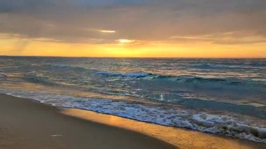 Gün batımında ve gündoğumunda deniz kıyısında mavi şeffaf suyla deniz dalgaları. Deniz dalgalarının yüzeyindeki güneş ışığının sarı yansıması. Koyu yağmur bulutları, ufuk çizgisi ve kumdaki deniz dalgaları..