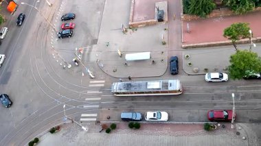 Asfaltlı sokak manzarası, toplu taşıma durağı, tramvay ve insanlar, hareket halindeki arabalar, tramvay rayları, yeşil ağaçlar, yürüyen insanlar ve yaz günü park halindeki arabalar. Açık havada. Yukarıdan görüntüle
