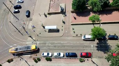 Yukarıdan şehir caddesi. Arabalar, tramvay rayları, hareket halindeki tramvay, park halindeki arabalar, yeşil ağaçlar ve güneşli bir yaz gününde şehir caddesinde yürüyen insanlar için bir asfalt yol. Üst Hava Görünümü.
