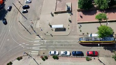 Yukarıdan şehir caddesi. Arabalar, tramvay rayları, hareket halindeki tramvay, park halindeki arabalar, yeşil ağaçlar ve güneşli bir yaz gününde şehir caddesinde yürüyen insanlar için bir asfalt yol. Üst Hava Görünümü.