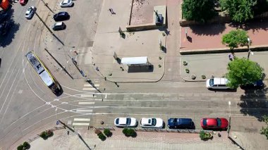 Yukarıdan şehir caddesi. Arabalar, tramvay rayları, hareket halindeki tramvay, park halindeki arabalar, yeşil ağaçlar ve güneşli bir yaz gününde şehir caddesinde yürüyen insanlar için bir asfalt yol. Üst Hava Görünümü.