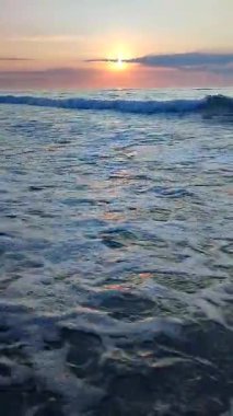Gün batımında ya da denizde şafakta. Okyanusta gün batımı ya da gün doğumu. Denizin üstünde güneş var. Güneş, gökyüzü, bulutlar, ufuk çizgisi, deniz, deniz dalgaları, kumlu sahil, sahil. Sabah ve akşam sahilde. Dikey