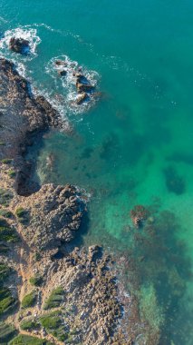 Temiz turkuaz suyun insansız hava aracı görüntüsü tenha bir kayalık sahil ve yeşil kıyı şeridiyle buluşuyor.