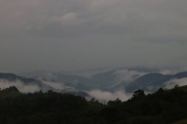 Yeşil ağaçlı yeşil dağ manzarası ve üzerinde mavi gökyüzü ve bulutlu çimenler.