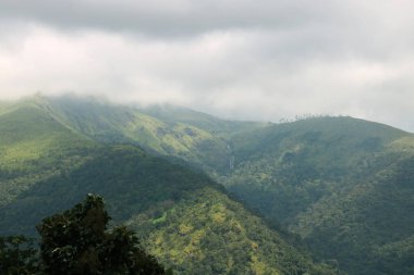 Yeşil ağaçlı yeşil dağ manzarası ve üzerinde mavi gökyüzü ve bulutlu çimenler.