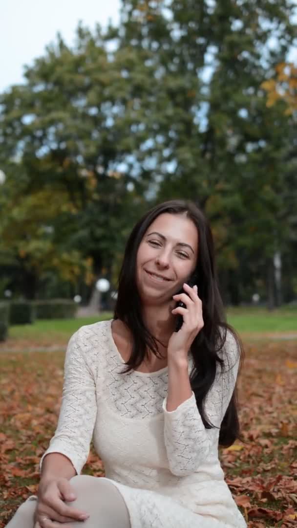 Joyeux belle fille assise dans un parc d'automne sur l'herbe et parlant sur un téléphone portable — Video