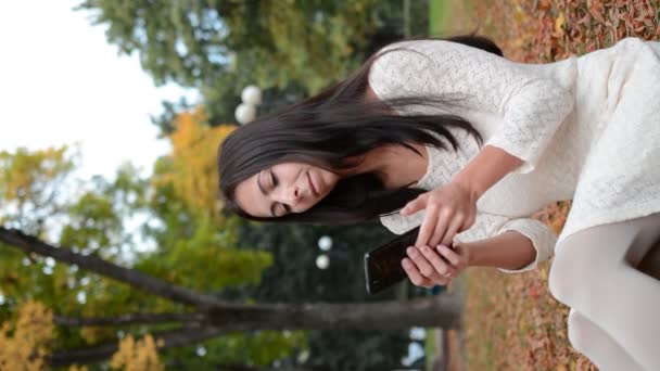 Uma menina morena se senta em um parque de outono na grama e escreve uma mensagem — Vídeo de Stock