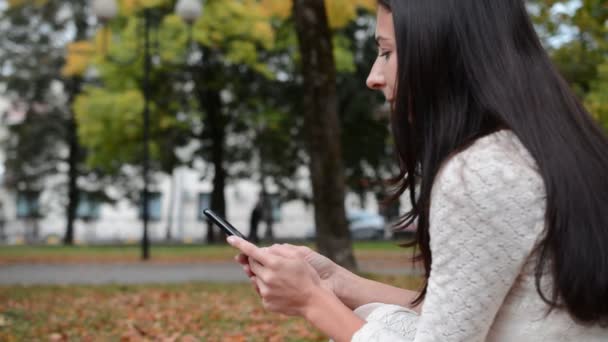 Vista lateral, una chica se sienta en un parque en el césped y escribe un mensaje — Vídeos de Stock