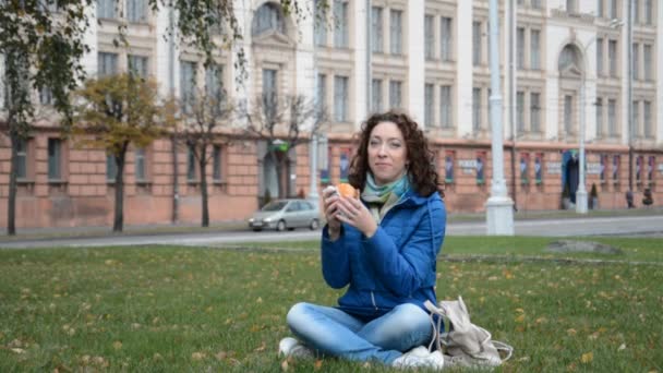 Mooi student meisje eet een hamburger of cheeseburger op het gazon — Stockvideo