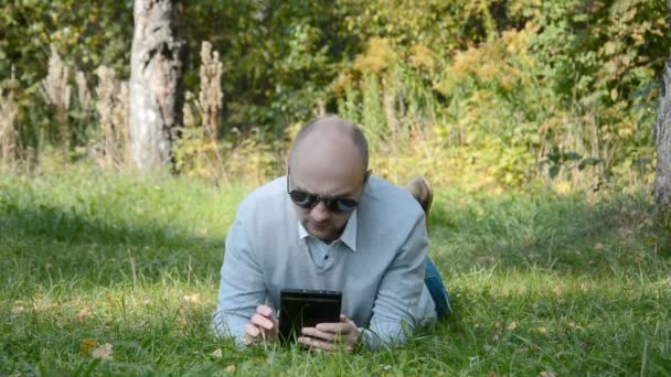 A man in glasses in the forest lies on the grass an communicates in a messenger on a tablet — Stock Video