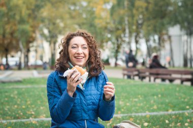Mavi ceketli kız, şehrinin caddesinde hamburger ya da çizburger yiyor.