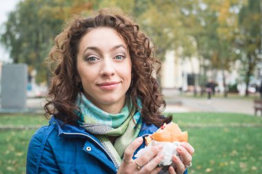 Mavi ceketli kız, şehrinin caddesinde hamburger ya da çizburger yiyor.