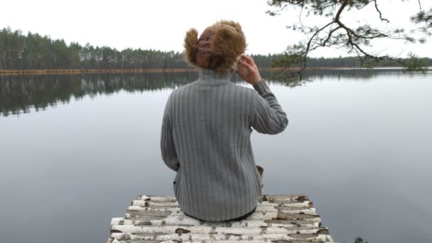 Vista trasera, un excursionista se sienta en la orilla de un lago y habla por teléfono. Tristeza y soledad, concepto de anhelo — Vídeos de Stock