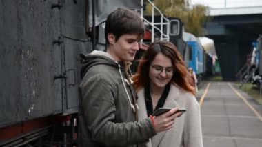 Bir adam ve bir kız tren istasyonunun platformunda dikiliyorlar ve birlikte video izliyorlar.
