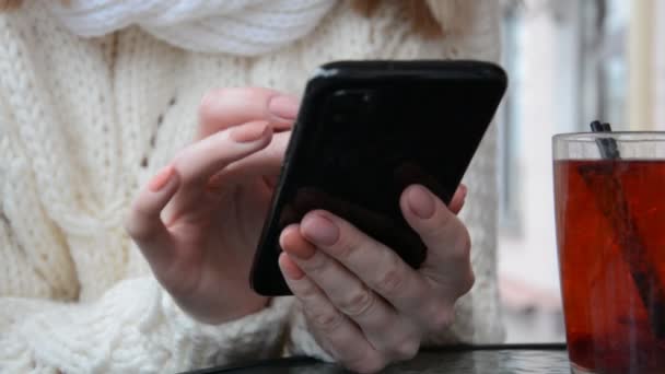 Закрыть руки деловой женщины в белом свитере, которая сидит и пользуется своим черным смартфоном — стоковое видео