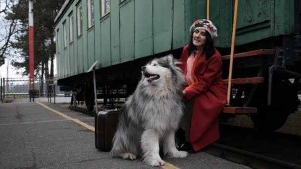 En kvinna i röd rock och en stor hund sitter på en gammal järnvägsplattform — Stockvideo