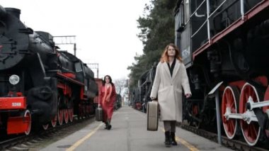 Bavullu iki kadın eski buhar lokomotiflerinin arasında bir tren platformu boyunca yürüyorlar.