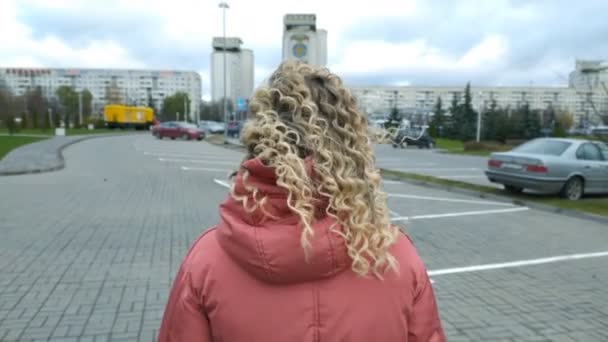 Kilátás az épület egy gyönyörű fiatal lány göndör haj sétál át egy autó parkoló a városban — Stock videók