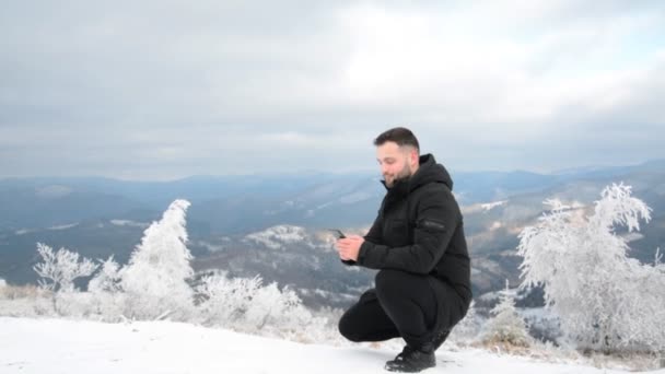 Przystojny facet siedzi na zaśnieżonej górze i pisze wiadomość przez telefon. — Wideo stockowe