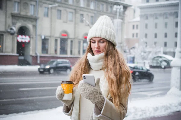 暖かい服にコーヒーと携帯電話を手にしたスタイリッシュな美しい女の子は 冬に街の通りで写真家のためにポーズ — ストック写真