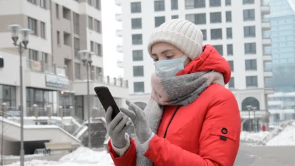 Una chica hipster con una chaqueta roja y una máscara protectora en su cara se para en su vecindario y escribe un mensaje — Vídeo de stock