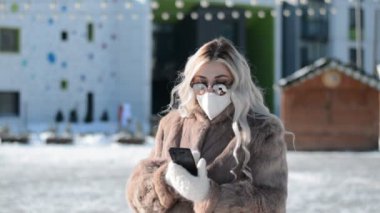 Koruyucu tıbbi maskeli ve güneş gözlüklü şık bir kadın kışın sokakta durup bir mesaj yazıyor.