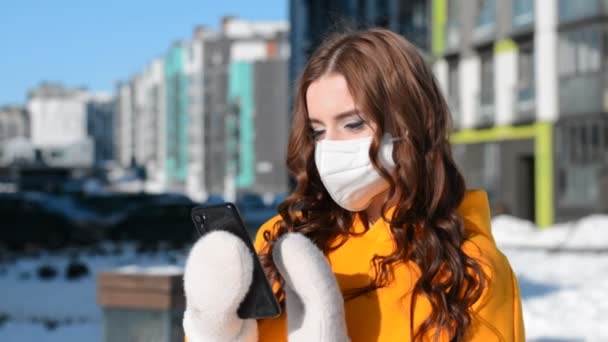 노란 후드를 쓴 러시아의 아름다운 소녀와 손에 전화기를 들고 야외 사진을 찍은 겨울의 보호용 마스크 — 비디오