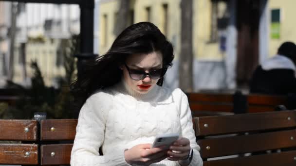 Bruneta hipster dívka komunikuje mobilním telefonem venku, zatímco sedí na lavičce ve městě — Stock video