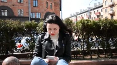 Otobüs durağında bankta oturan beyaz bir kadın internetten mesaj atmak için akıllı telefon kullanıyor.