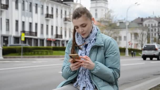 Egy gyönyörű fiatal lány fülhallgatóban zenét hallgat, és üzenetet ír egy okostelefonra a város utcáin. — Stock videók