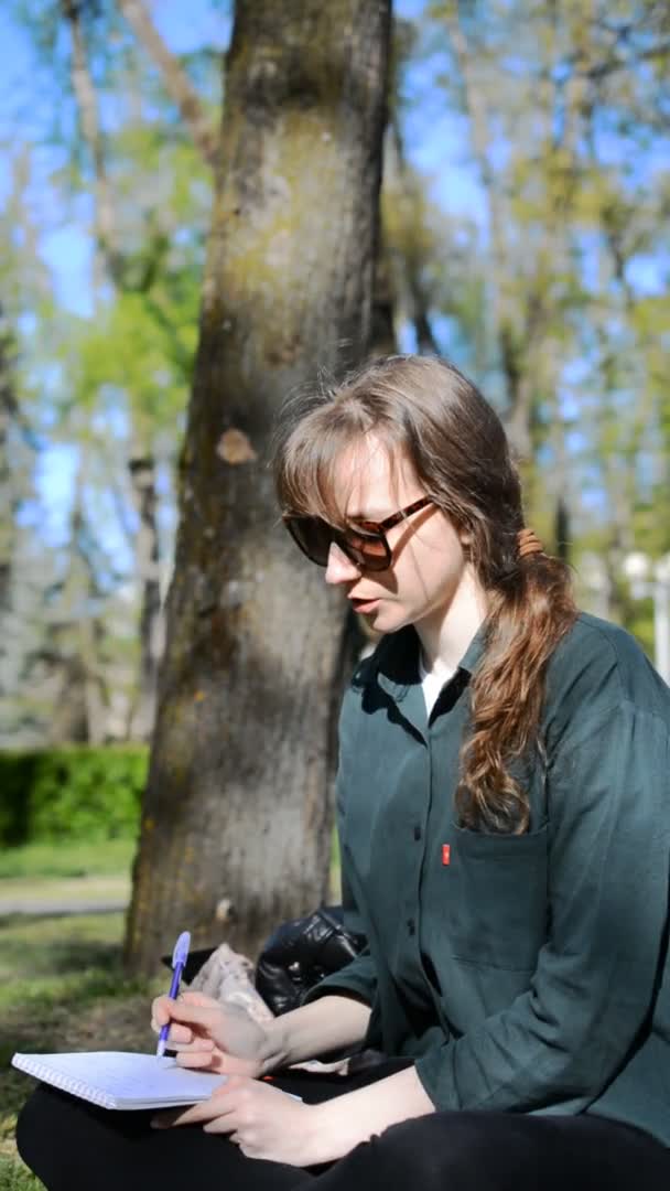一位年轻的高加索女学生坐在公园的草坪上，在笔记本上写字 — 图库视频影像