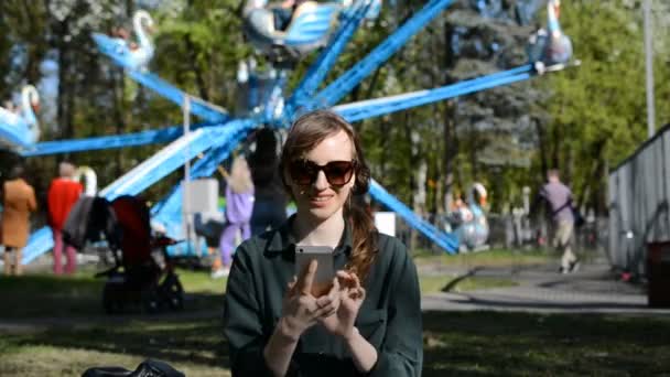 Een mooi jong meisje zit op een gazon in een park op een mooie zonnige zomerdag en schrijft een bericht op haar smartphone — Stockvideo