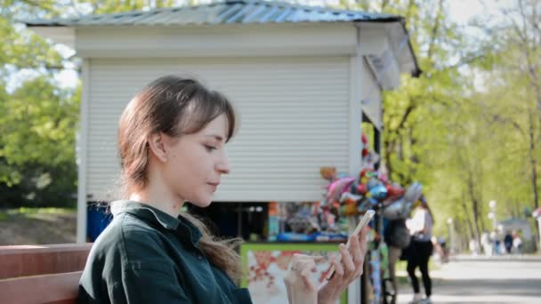 Bella giovane donna adulta seduta su una panchina nel parco della città e utilizzando il suo smartphone — Video Stock