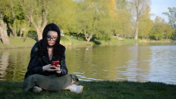 Piękna młoda brunetka w okularach siedzi na brzegu miejskiego jeziora i pisze wiadomość — Wideo stockowe